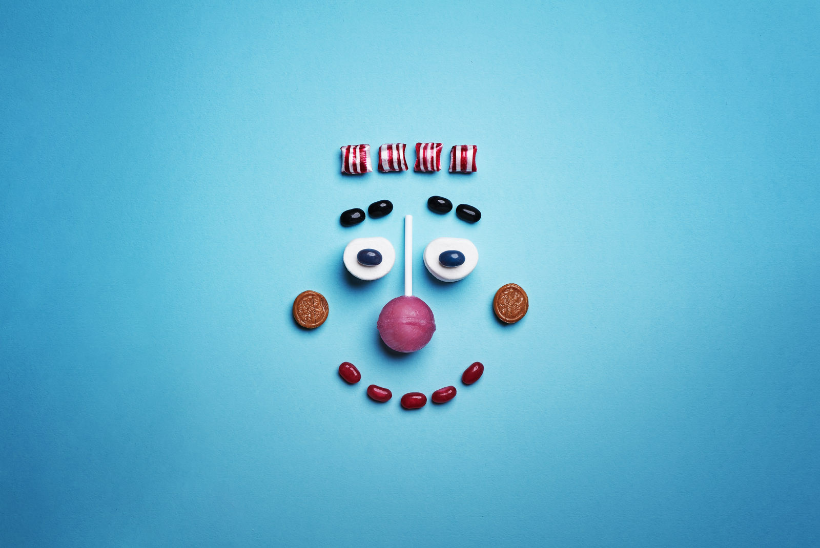 Ein lachendes Gesicht, bestehend aus Drops, Bonbons und einem Lutscher, fotografiert auf blauem Untergrund.